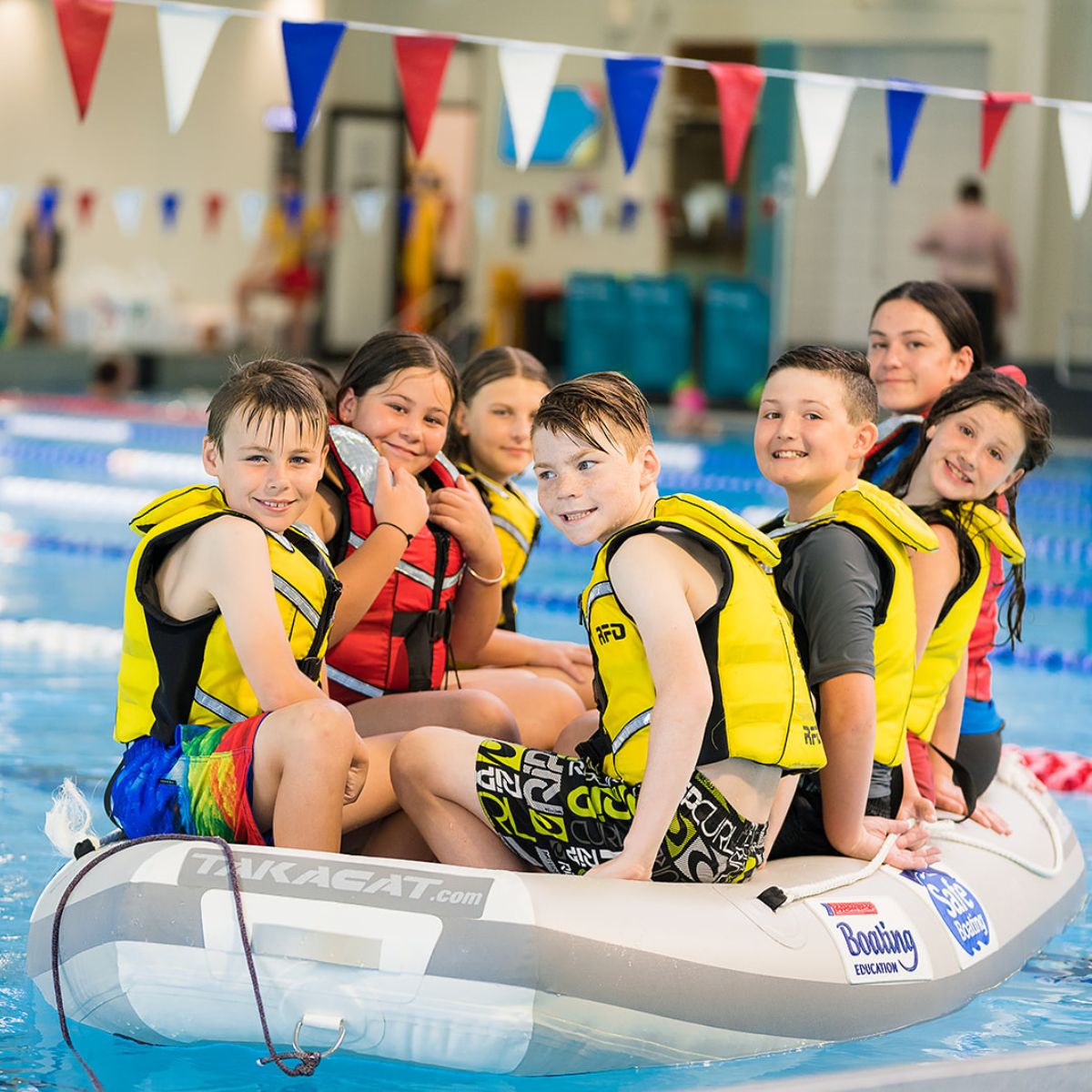 A group of smiling boys and girls in a flotation raft wearing yellow life jackets at Christchurch Recreation and Sport pool. 
