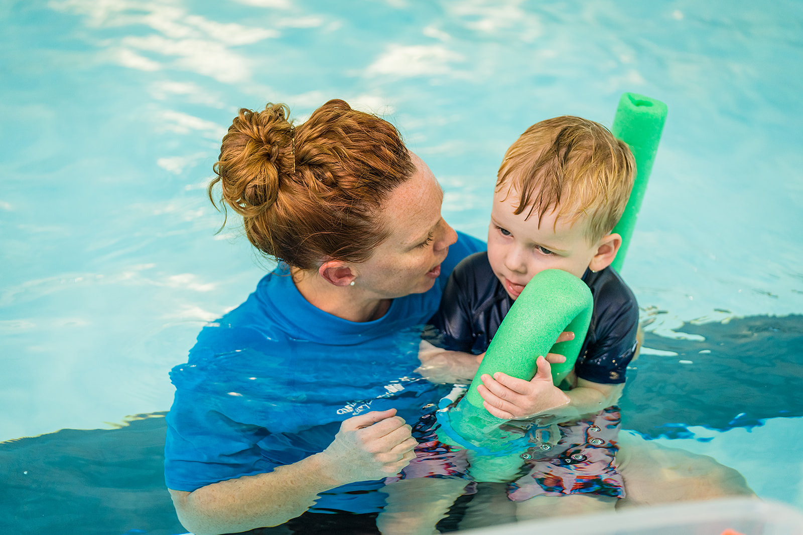 A little boy wearing a blue swimsuit floating in the pool holding onto a floatie while a female instructor holds the child in her arms.
