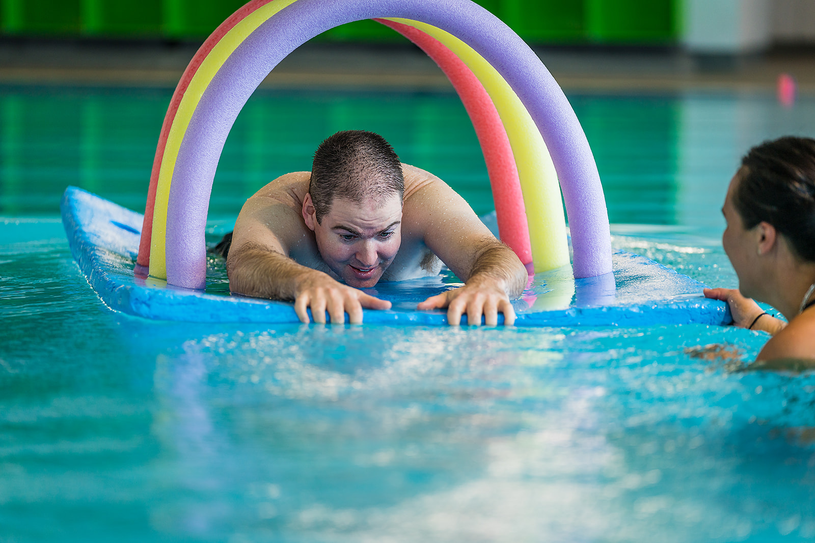 A smiling man playing on a floating blue pool mat during a Sensory Swimming class at Christchurch Recreation and Sport pool.