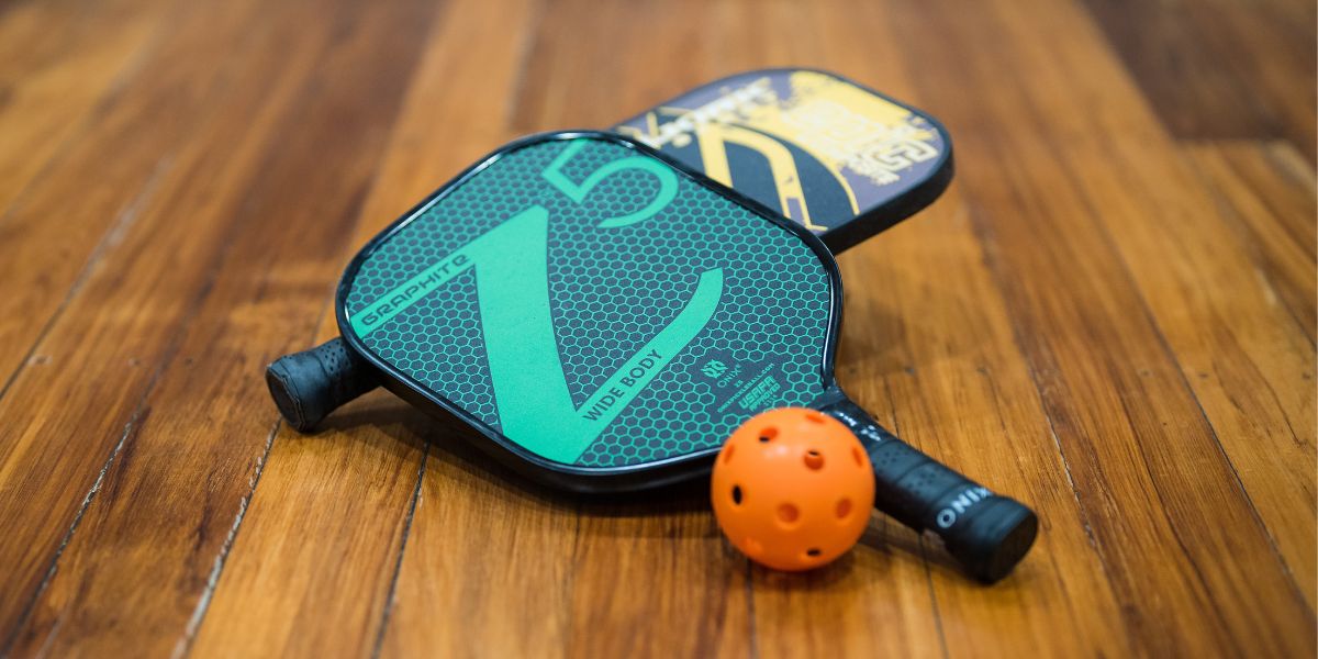 Two pickleball rackets, blue and yellow placed on the court with an orange wiffle ball. 