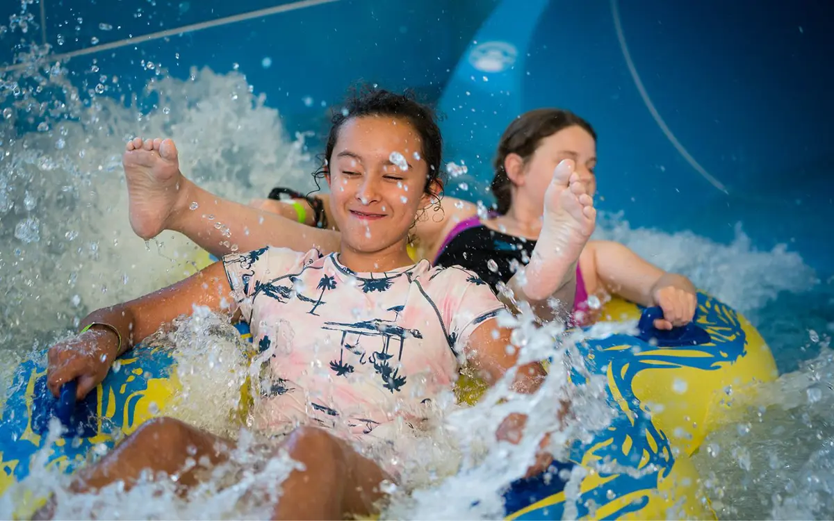 Two smiling girls sliding down the blue hydroslide at QEll Recreation and Sport Centre in a yellow tandem raft.