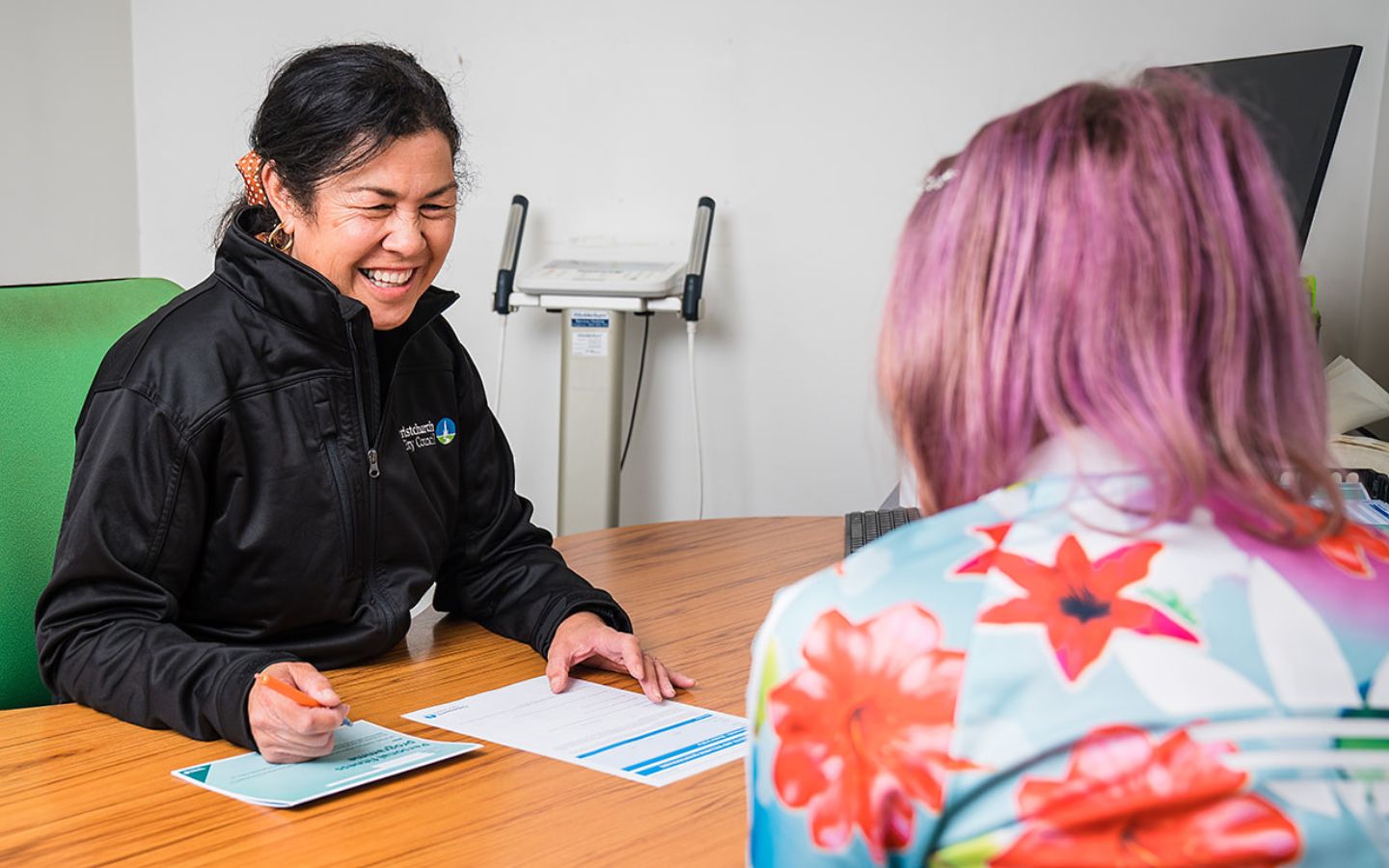 A smiling female personal trainer in a black staff uniform sitting across a desk from a female client at Christchurch Recreation and Sport Centre.