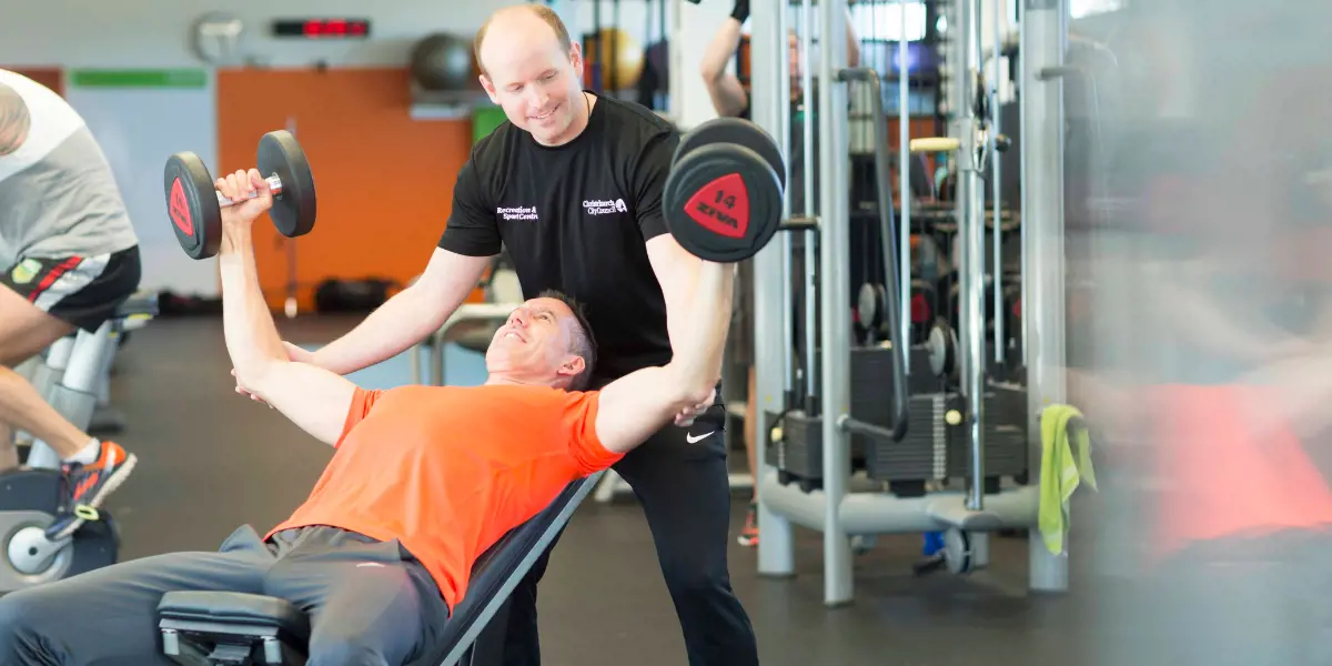 A male personal trainer in a black staff uniform assisting a male client in orange with a shoulder press at Christchurch Recreation and Sport Centre.