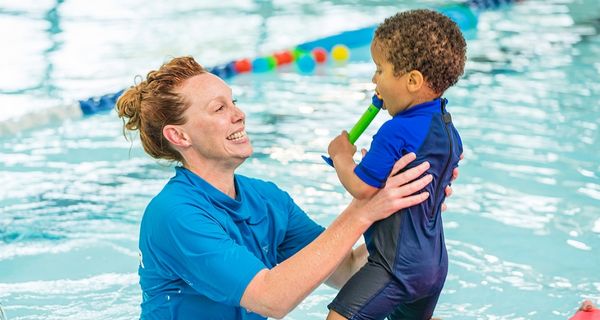A little boy wearing a blue swimsuit is jumping into the arms of a smiling female swim instructor as she stands in the pool. 