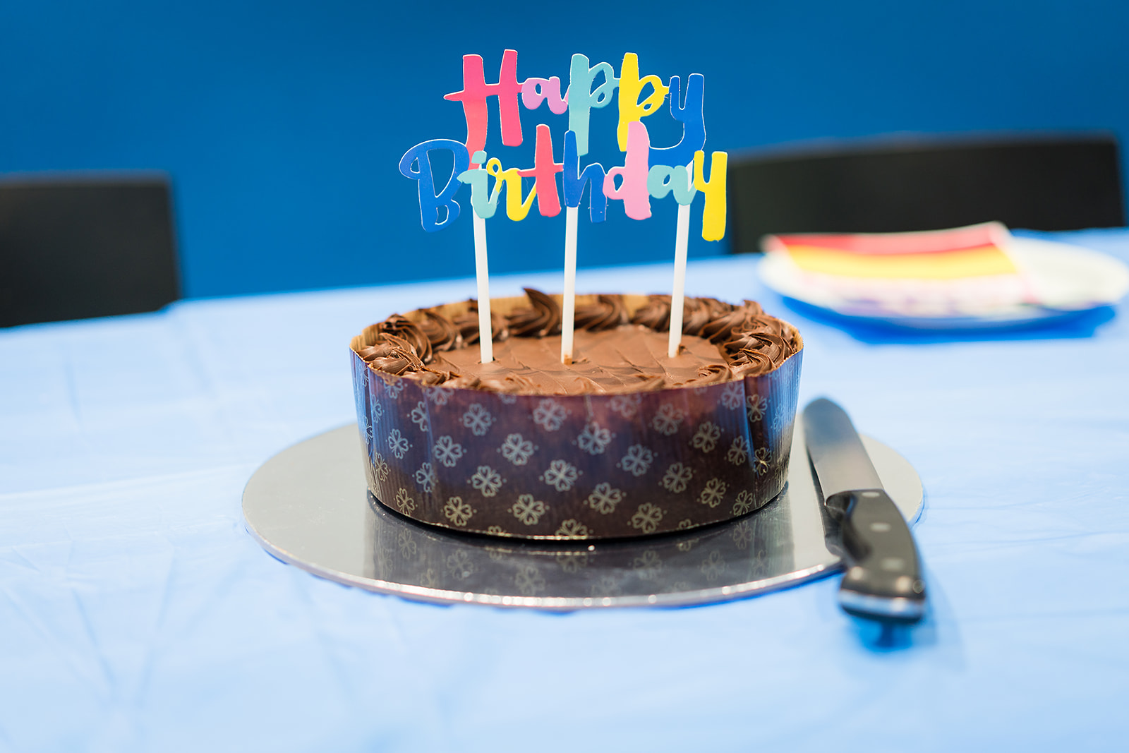 A chocolate cake with a Happy Birthday sign on the top. Sitting on a blue table. 