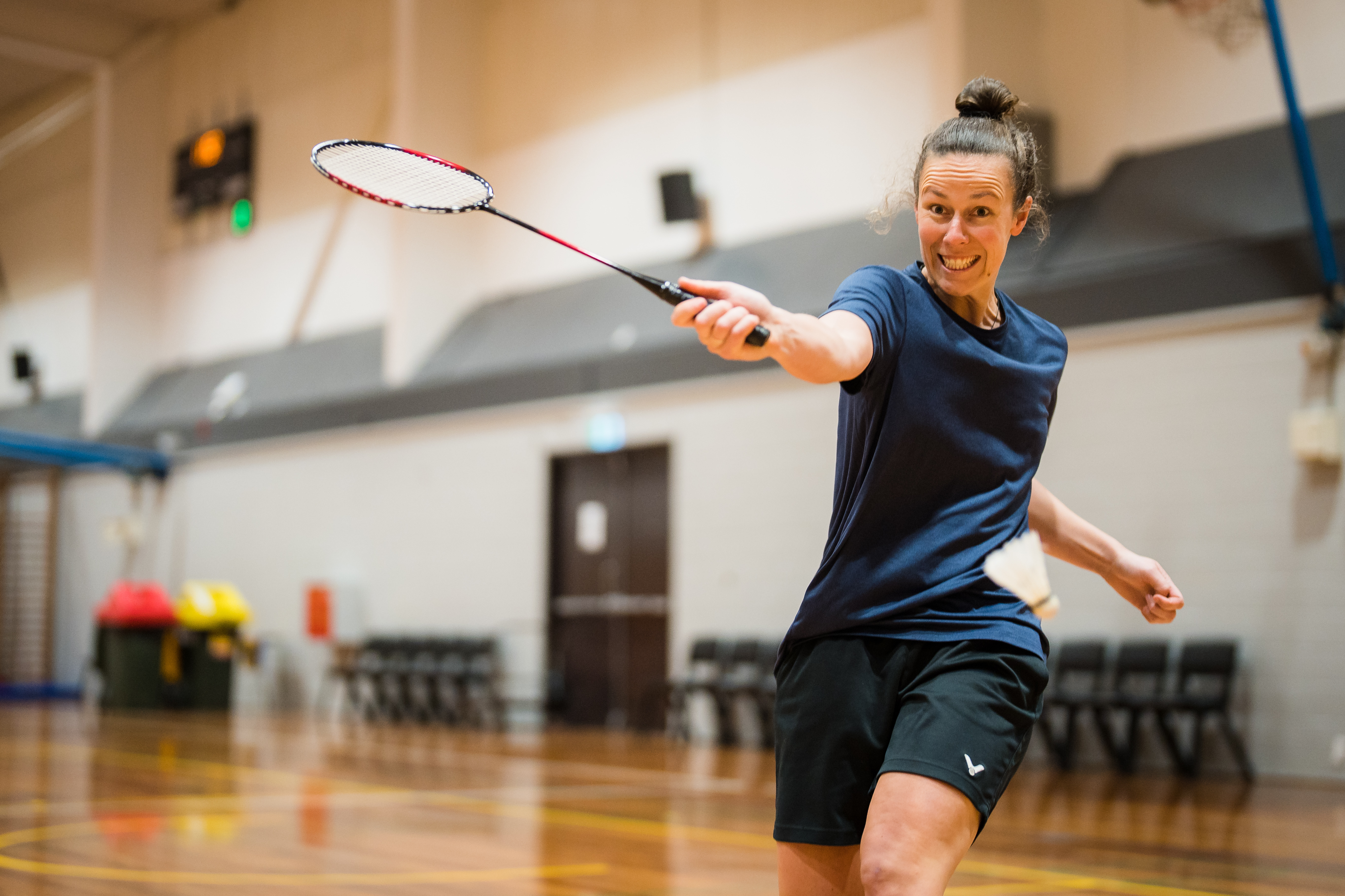 A woman smiling while holding a badminton racket hitting a white birdie at Christchurch Recreation and Sport. 
