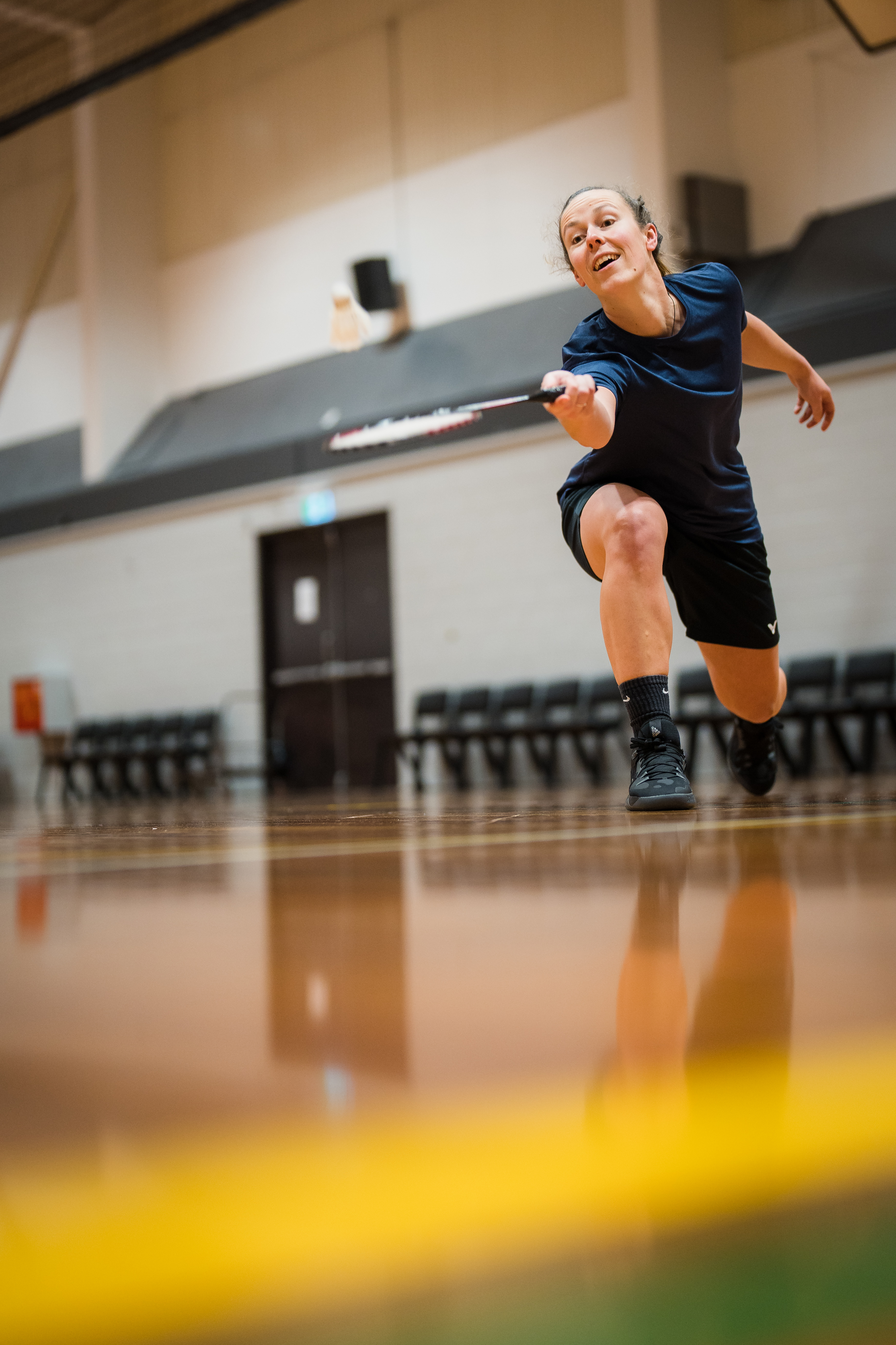 A woman smiling while holding a badminton racket hitting a white birdie at Christchurch Recreation and Sport. 