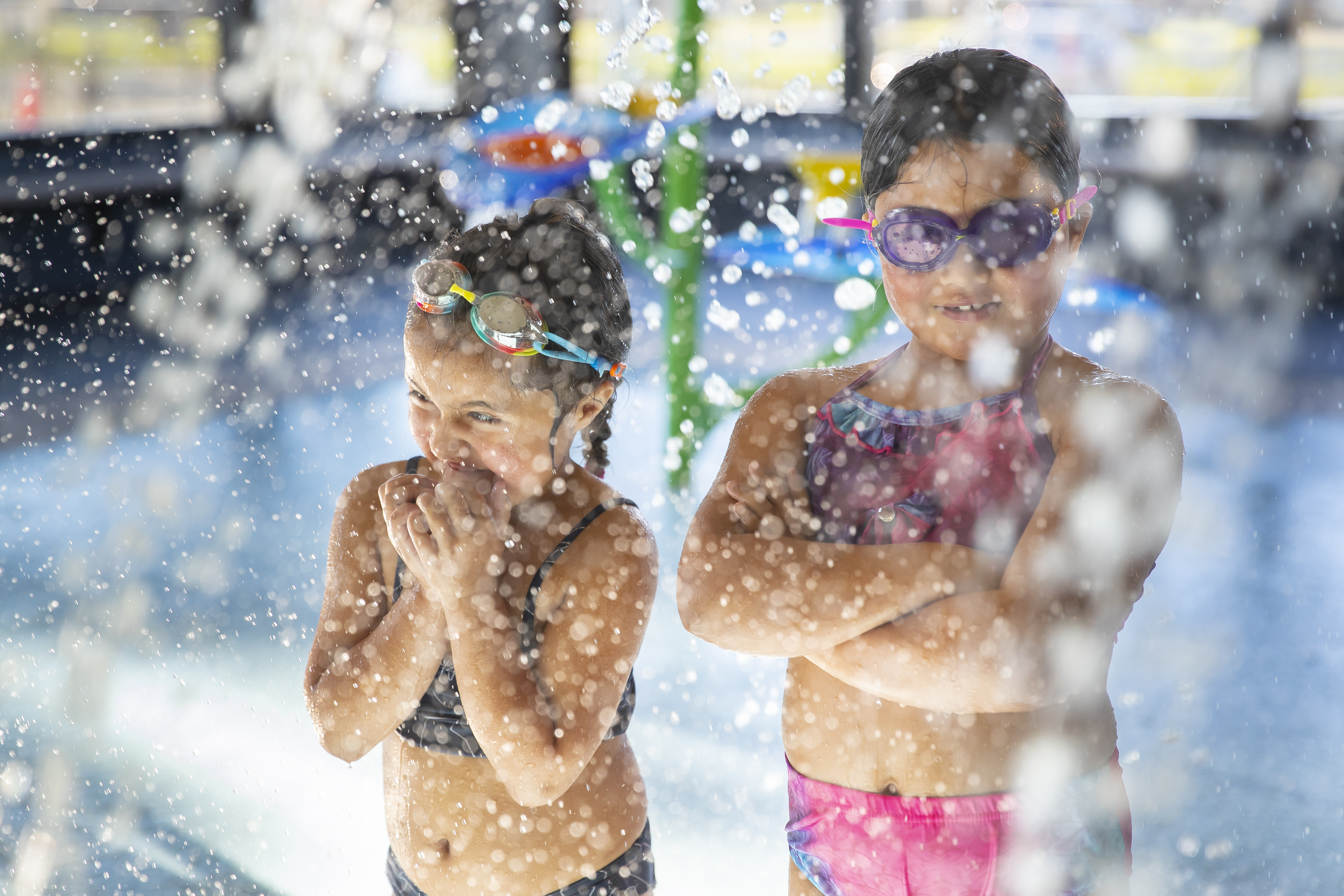 Two girls with their arms crossed wearing goggles as they stand under a children's pool waterfall at Matatiki Hornby Centre. 
