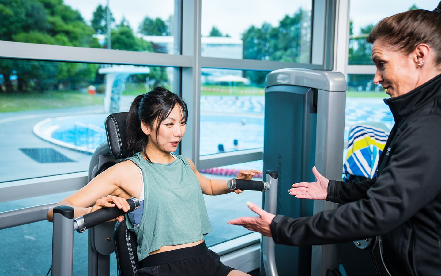 A female personal trainer in a black staff uniform assisting a female client with a shoulder press at Christchurch Recreation and Sport Centre.