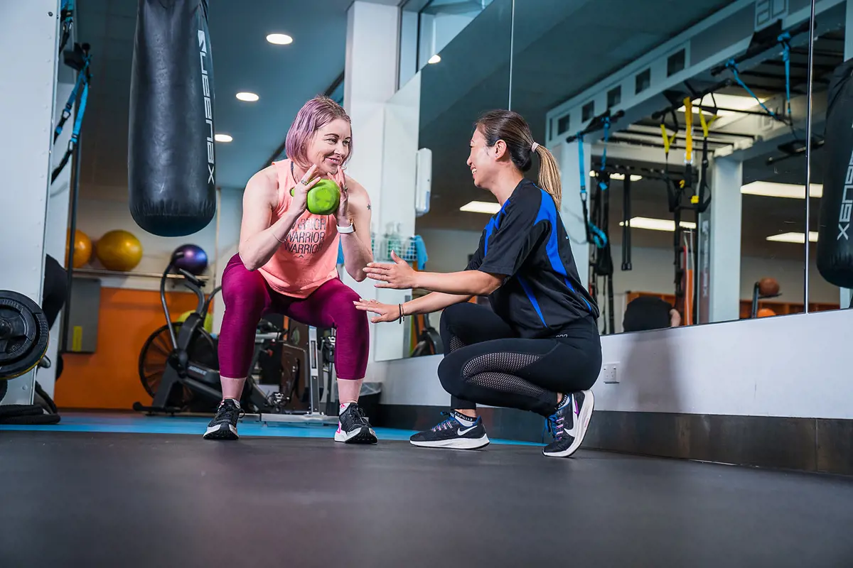 A female personal trainer assisting a woman in pink through a kettle bell workout at Christchurch Recreation and Sport Centre.